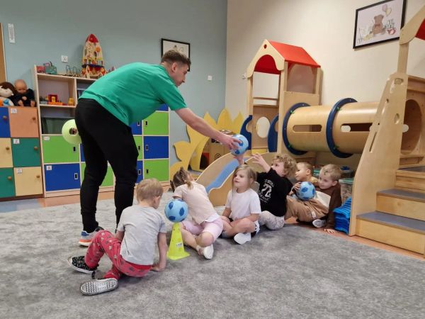 Trener wręcza piłkę dzieciom siedzącym w rzędzie podczas zajęć sportowych Sportuś
